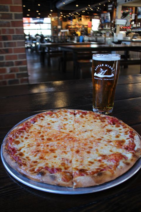 A cheese pizza and a pint of beer with the Little Miami Brewing Company logo, inside a brewery with a cozy atmosphere.