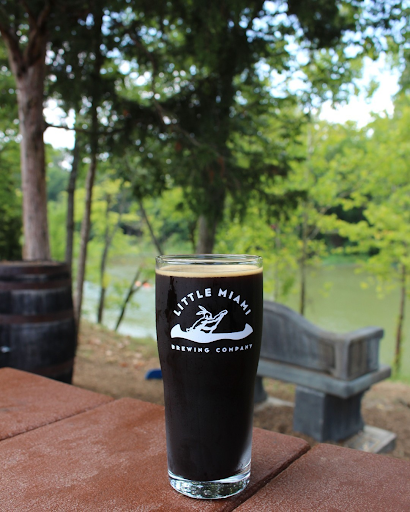 A dark beer in a glass with the Little Miami Brewing Company logo, set against lush greenery by the river.
