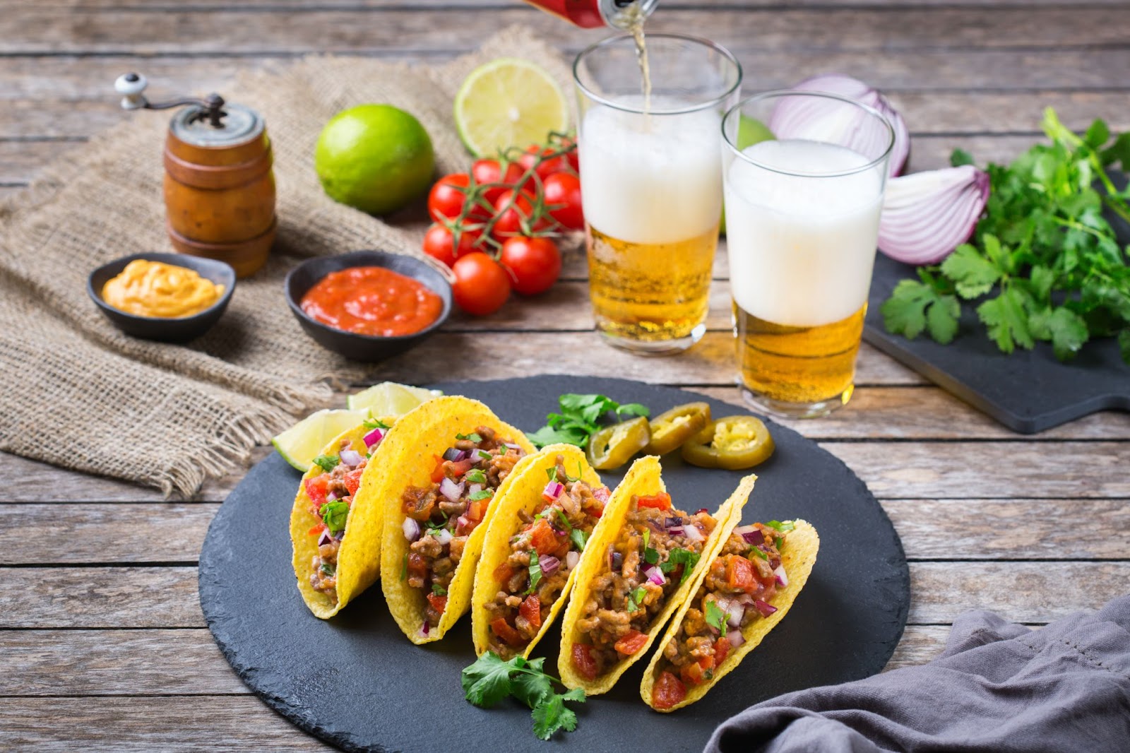 A vibrant spread of tacos A vibrant spread of tacos filled with colorful toppings on a slate platter, accompanied by fresh ingredients, sauces, and glasses of frothy beer being poured.