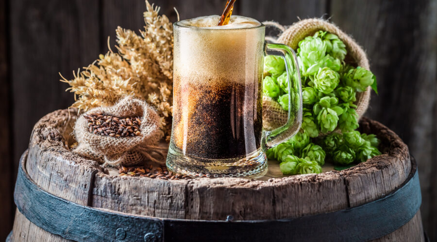 Dark beer being poured into a glass mug on a wooden barrel with hops and barley in the background.